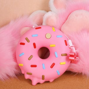 May include: A pink donut-shaped purse with colourful sprinkles and a strap. The purse has a melted frosting design on the bottom and a small button on top. The background is a soft, pink, furry texture.