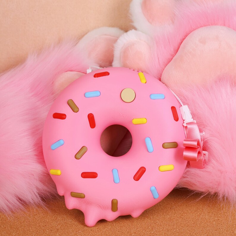 May include: A pink donut-shaped purse with colourful sprinkles and a strap. The purse has a melted frosting design on the bottom and a small button on top. The background is a soft, pink, furry texture.