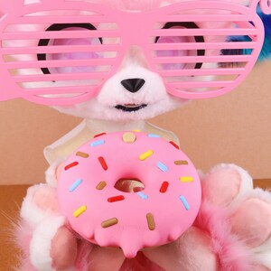 May include: A soft pink toy animal wearing pink shutter shades, holding a pink donut with colourful sprinkles. The toy has white fur and a black nose. The donut is the central focus, with a hole in the middle.