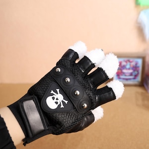 Puede incluir: Guantes negros sin dedos con un dise&ntilde;o de calavera y huesos cruzados y ribete de piel blanca. Los guantes tienen tachuelas met&aacute;licas en la parte posterior de la mano.