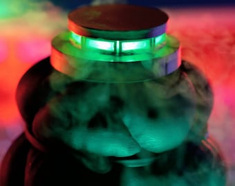 May include: A black and silver smoke machine with a green light illuminating the top. The machine is surrounded by a cloud of white smoke.