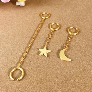 May include: Gold-colored ear cuffs with chain accents. One cuff features a long chain, while the others have a star and crescent moon charm. The jewelry is displayed on a textured, tan surface.