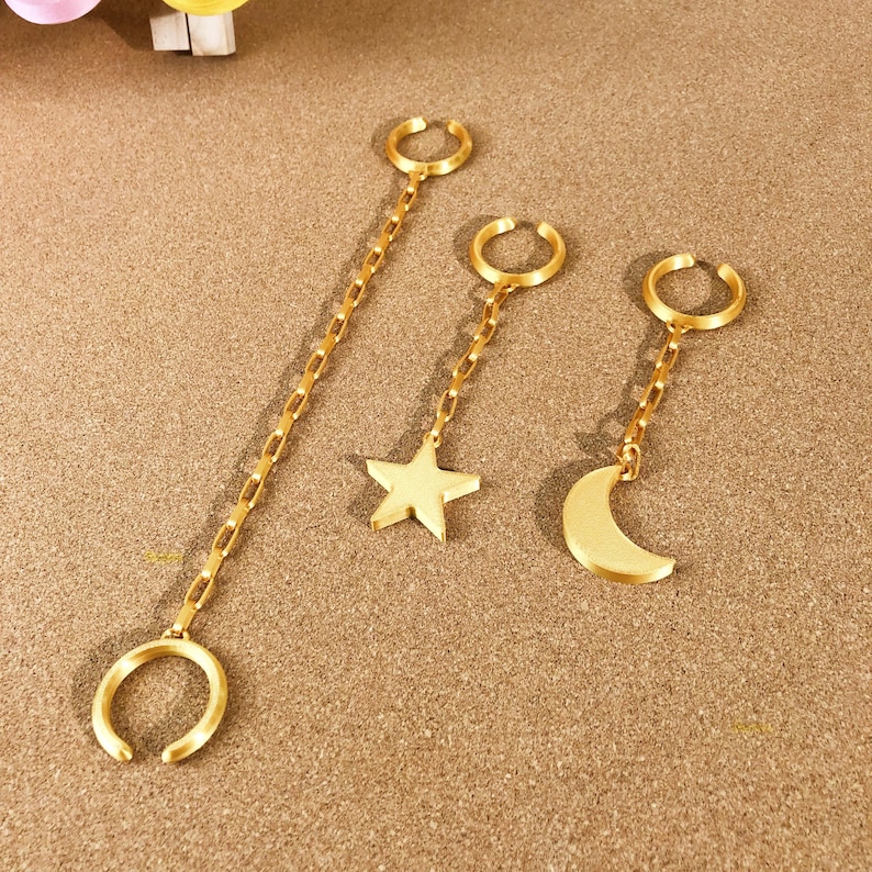 May include: Gold-colored ear cuffs with chain accents. One cuff features a long chain, while the others have a star and crescent moon charm. The jewelry is displayed on a textured, tan surface.
