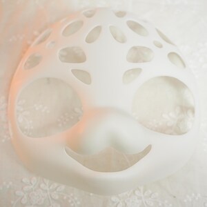 Puede incluir: M&aacute;scara facial de pl&aacute;stico blanco con recortes para los ojos, la nariz y la boca. La m&aacute;scara tiene un dise&ntilde;o caprichoso con una boca sonriente y un patr&oacute;n de peque&ntilde;os c&iacute;rculos alrededor de los ojos.