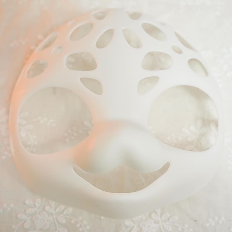 Puede incluir: M&aacute;scara facial de pl&aacute;stico blanco con recortes para los ojos, la nariz y la boca. La m&aacute;scara tiene un dise&ntilde;o caprichoso con una boca sonriente y un patr&oacute;n de peque&ntilde;os c&iacute;rculos alrededor de los ojos.