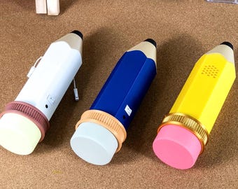 Puede incluir: Tres altavoces Bluetooth con forma de lápiz en blanco, azul y amarillo con una goma de borrar marrón en la parte inferior.