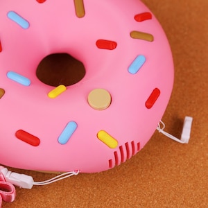 May include: A pink donut-shaped object with colourful sprinkles. The donut has a brown centre and a beige button on the side. It has a white cord and a pink and white strap attached.