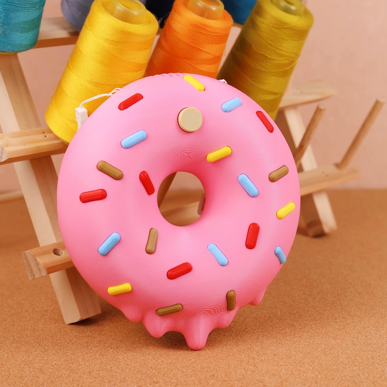 May include: A pink donut-shaped purse with colourful sprinkles. The purse has a round shape with a hole in the centre and a button closure. The sprinkles are red, blue, yellow, and brown. The purse is on a light brown surface.