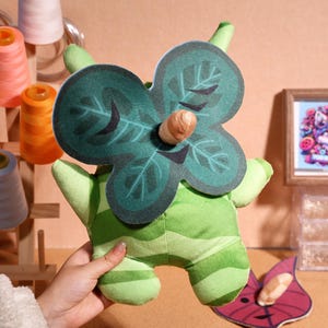 Può includere: Giocattolo di peluche verde con un design a foglia e un corno marrone. Il giocattolo ha un viso sorridente ed è fatto di tessuto morbido e peluche.