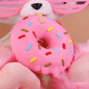 May include: A pink donut with colourful sprinkles, including red, yellow, blue, and brown, is the focal point. The donut has a hole in the centre and a glossy finish. The background includes a plush pink surface and pink-framed glasses.