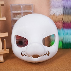 Könnte beinhalten: Eine weiße, leere Maske im Cartoon-Stil mit großen Augenöffnungen, einer markanten Nase und einem gezackten Mund. Die Maske besteht aus einem glatten Material und ist für Bastelarbeiten oder Cosplay konzipiert. Bastelbedarf ist im Hintergrund zu sehen.