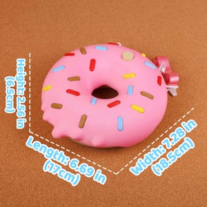 May include: A pink donut with pink icing and colourful sprinkles. The donut measures 17cm in length, 18.5cm in width, and 6.5cm in height. A small pink bow is attached to the side.