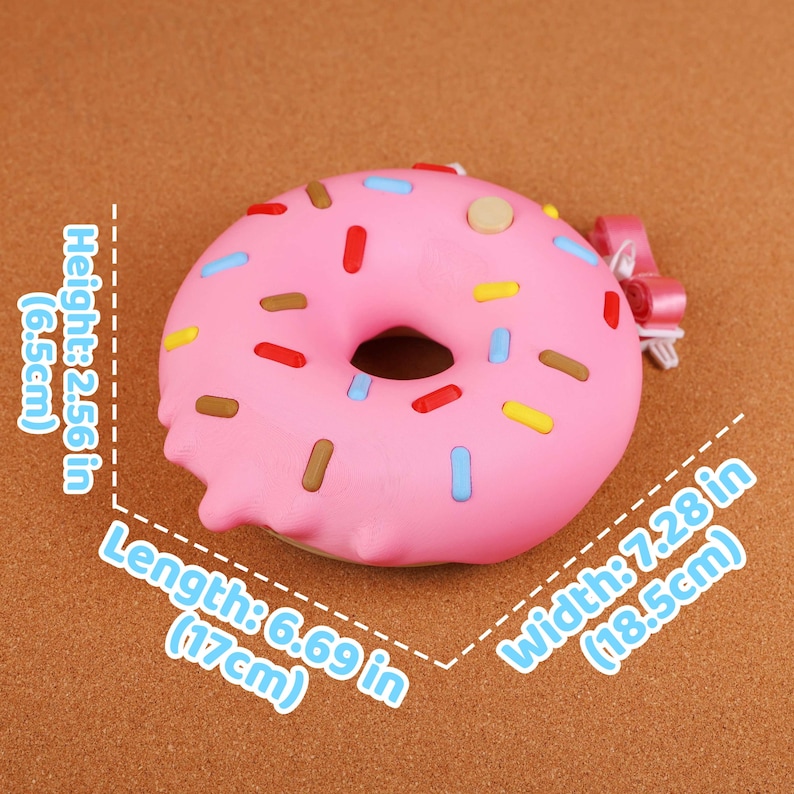 May include: A pink donut with pink icing and colourful sprinkles. The donut measures 17cm in length, 18.5cm in width, and 6.5cm in height. A small pink bow is attached to the side.