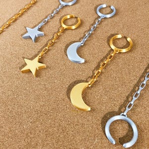 May include: Ear cuff earrings in gold and silver tones. The earrings feature star and crescent moon charms, connected to the ear cuff by a chain. The earrings are displayed on a brown surface.