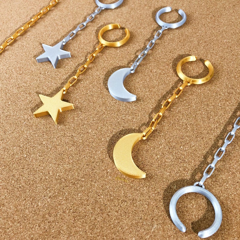 May include: Ear cuff earrings in gold and silver tones. The earrings feature star and crescent moon charms, connected to the ear cuff by a chain. The earrings are displayed on a brown surface.