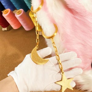 May include: A gold-colored chain with a crescent moon and star charm. The chain is attached to two gold rings. The background includes pink and white faux fur and spools of thread in various colors.