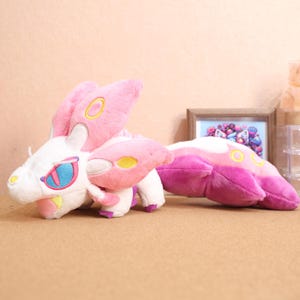 May include: A pink and white plush toy with yellow accents. The toy has a long tail and large, pointed ears. It has a white face with a blue eye and a pink mouth.