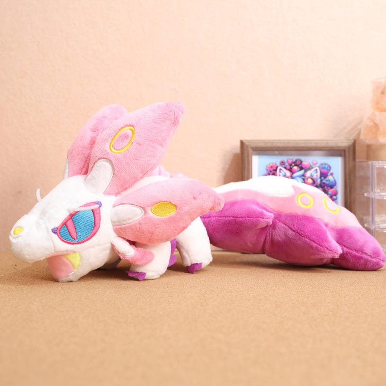 May include: A pink and white plush toy with yellow accents. The toy has a long tail and large, pointed ears. It has a white face with a blue eye and a pink mouth.