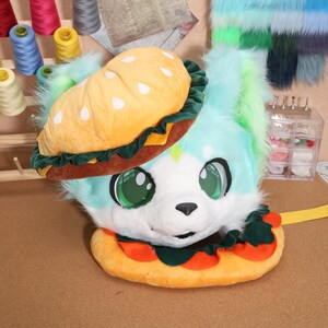 May include: A plush, furry headpiece in the shape of a cartoon animal with large green eyes, wearing a burger hat and a burger-shaped collar. The burger hat is yellow with white sesame seed details, and the collar has red tomato accents.