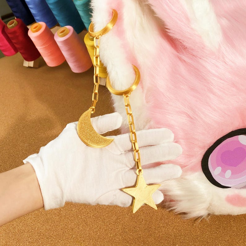 May include: Gold-colored crescent moon and star charm hanging from a chain. The charm is held by a gloved hand, with a pink and white furry texture in the background. Spools of thread are visible in the background.