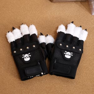 Puede incluir: Un par de guantes negros sin dedos con piel sint&eacute;tica blanca en la parte posterior de la mano y los dedos. Los guantes tienen un dise&ntilde;o de calavera y huesos cruzados en la palma y tachuelas plateadas.