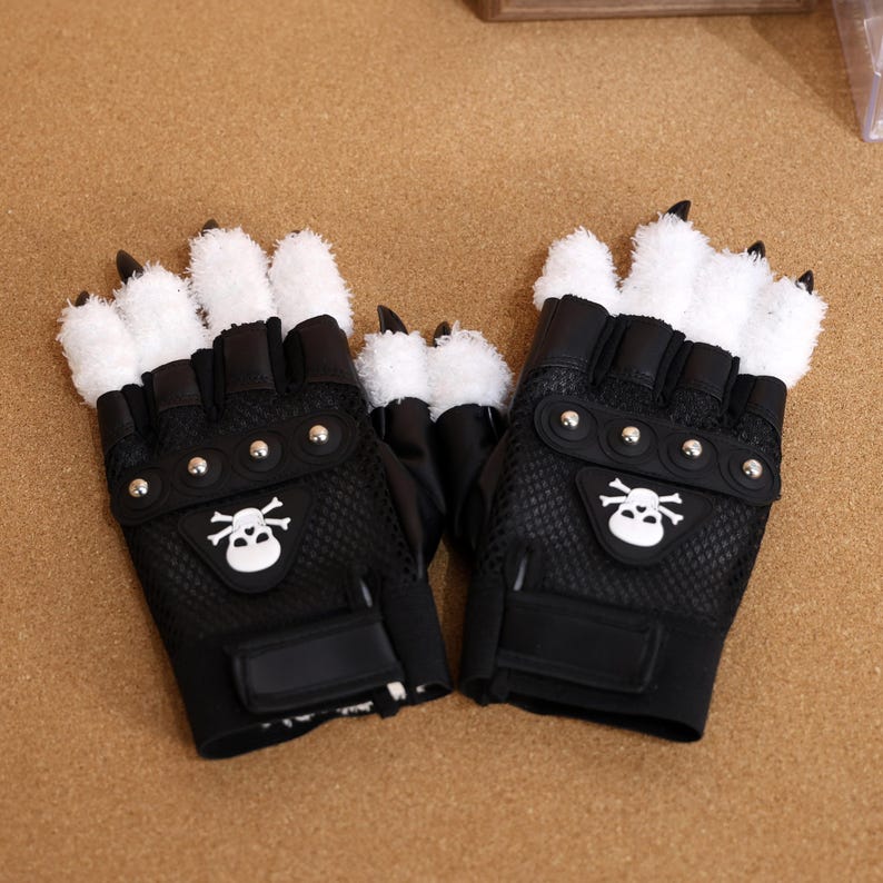 Puede incluir: Un par de guantes negros sin dedos con piel sint&eacute;tica blanca en la parte posterior de la mano y los dedos. Los guantes tienen un dise&ntilde;o de calavera y huesos cruzados en la palma y tachuelas plateadas.