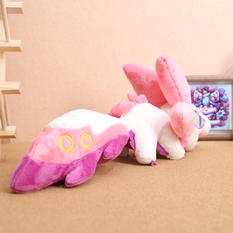 May include: Pink and white plush toy with purple accents. The toy has a long tail with three gold rings and large pink ears.