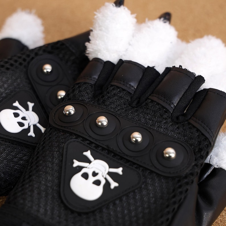 Puede incluir: Guantes negros sin dedos con un dise&ntilde;o de calavera y huesos cruzados blancos. Los guantes tienen una tela de malla negra y tachuelas plateadas.