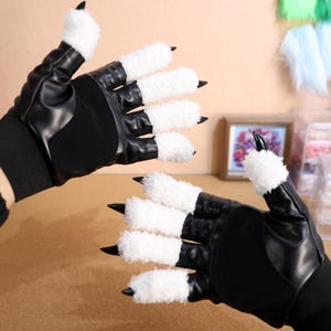 Puede incluir: Un par de guantes de cuero negro con garras blancas y peludas. Los guantes tienen un acabado de cuero texturizado y est&aacute;n dise&ntilde;ados para parecer patas de animales.