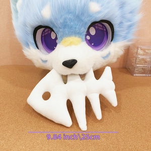 Può includere: Una testa di animale in peluche, blu e bianca, con grandi occhi viola e un naso nero. Uno scheletro di pesce bianco è tenuto in bocca. L'articolo misura 25 cm di larghezza.