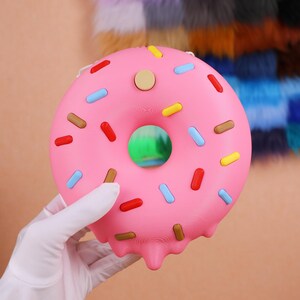 May include: A pink donut with colourful sprinkles and a light brown centre. The donut has a hole in the middle and a dripping effect on the bottom. The sprinkles are red, blue, yellow, and brown.