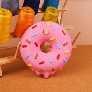 May include: A pink donut-shaped object with colourful sprinkles and a light brown centre. The donut has a melted glaze effect at the bottom. Spools of thread in various colours are in the background.