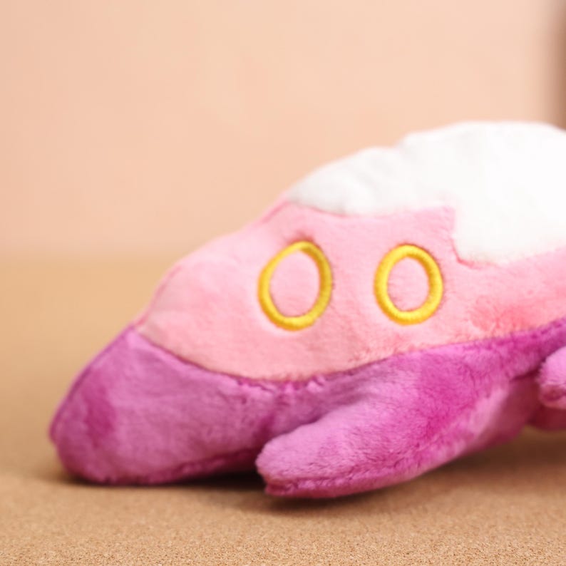 May include: Pink and purple plush toy shaped like a spaceship with two yellow embroidered circles on the front.