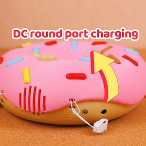 May include: A pink donut-shaped device with colourful sprinkles and the text "DC round port charging." The device has a yellow arrow pointing towards a charging port. The bottom half is tan.