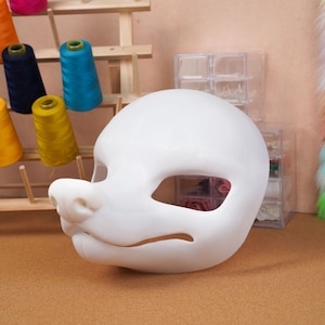 May include: A white, blank fursuit mask base with eye and mouth openings. The mask has a rounded shape with a prominent snout and is likely for a custom-made costume. Colorful spools of thread are visible in the background.