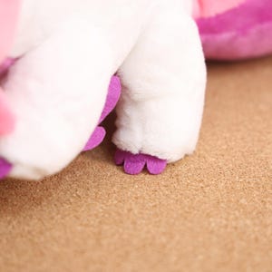 May include: Close-up of a white plush toy with purple feet. The toy is lying on a brown surface.