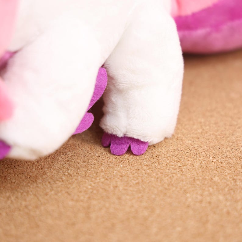 May include: Close-up of a white plush toy with purple feet. The toy is lying on a brown surface.