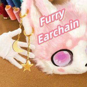 May include: Gold-colored ear chains with a crescent moon and star charm, displayed near a pink and white furry mask. The text "Furry Earchain" is visible. The background includes spools of thread.