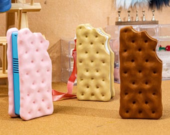 Puede incluir: Tres bolsos rectangulares con forma de sándwiches de helado, cada uno con una superficie texturizada con hoyuelos. Un bolso es rosa claro con un lado azul, otro es beige y otro es marrón oscuro. Cada bolso tiene un recorte en forma de mordisco.