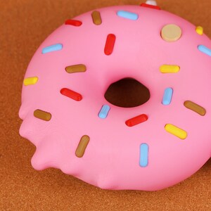 May include: A close-up of a pink donut with colourful sprinkles. The donut has a hole in the centre and is decorated with red, blue, yellow, and brown sprinkles. The donut is on a brown surface.