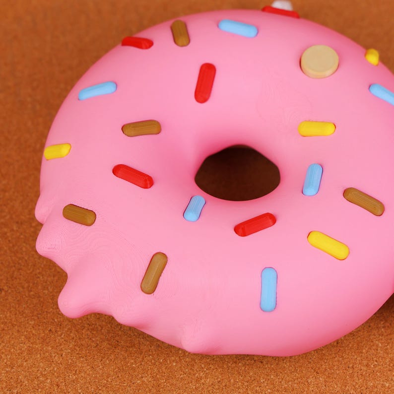 May include: A close-up of a pink donut with colourful sprinkles. The donut has a hole in the centre and is decorated with red, blue, yellow, and brown sprinkles. The donut is on a brown surface.
