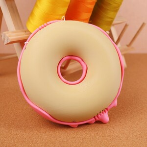 May include: A beige donut-shaped object with a pink outline and a pink centre. The donut has a pink frosting drip on the bottom. The background includes spools of yellow, orange, and green thread.