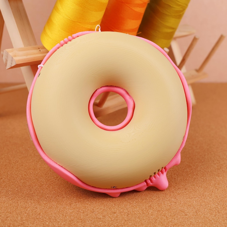 May include: A beige donut-shaped object with a pink outline and a pink centre. The donut has a pink frosting drip on the bottom. The background includes spools of yellow, orange, and green thread.