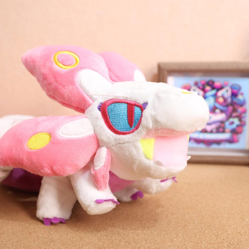 May include: Pink and white plush toy with large wings, yellow and white spots, and red and blue eyes. The toy has a large open mouth with yellow teeth.