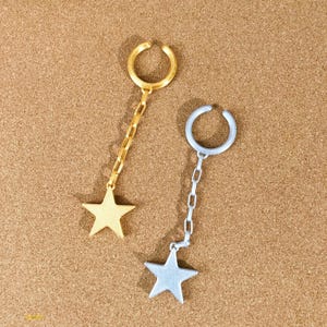 May include: Two ear cuffs, one gold-toned and one silver-toned, each with a dangling star charm. The cuffs have a curved design to fit around the ear. The star charms are connected to the cuffs by a chain.