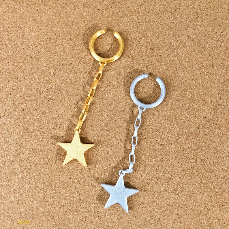May include: Two ear cuffs, one gold-toned and one silver-toned, each with a dangling star charm. The cuffs have a curved design to fit around the ear. The star charms are connected to the cuffs by a chain.