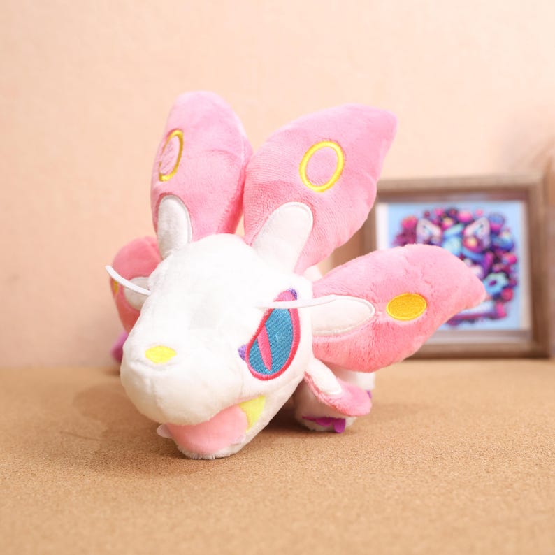 May include: A pink and white plush toy with large, butterfly-like wings. The toy has a yellow and pink face with a single, large, blue and pink eye.