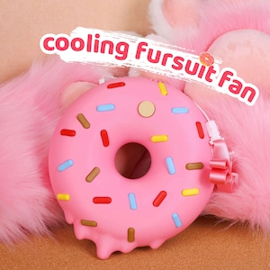 May include: A pink donut-shaped cooling fan with colourful sprinkles and the text "cooling Fursuit fan". The fan has a strap and a button. The background is a fluffy pink texture.