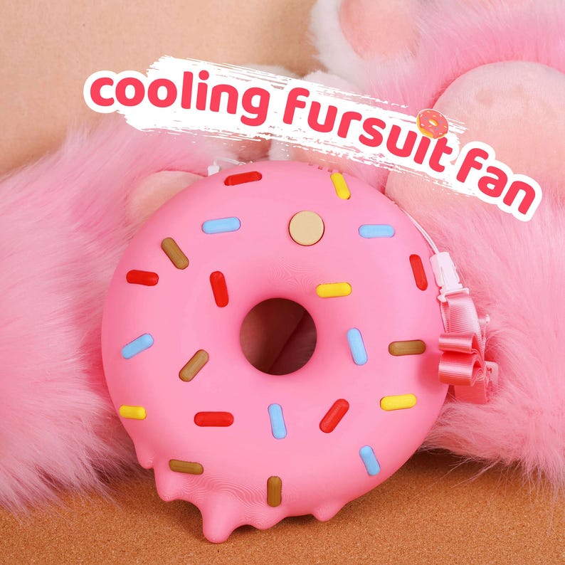 May include: A pink donut-shaped cooling fan with colourful sprinkles and the text "cooling Fursuit fan". The fan has a strap and a button. The background is a fluffy pink texture.