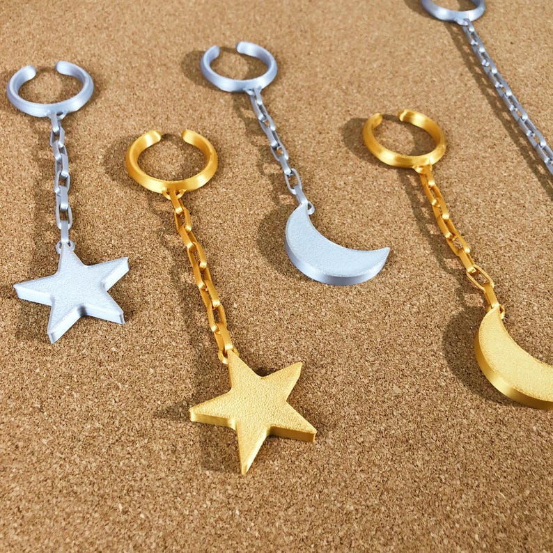 May include: Ear cuff jewelry with celestial designs. The image shows several ear cuffs with a silver star, a gold star, and silver and gold crescent moons. Each cuff has a chain connecting the ear cuff to the celestial charm.